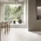 Minimalist cafe interior with wooden tables, chairs, and a large potted tree by expansive windows overlooking greenery.