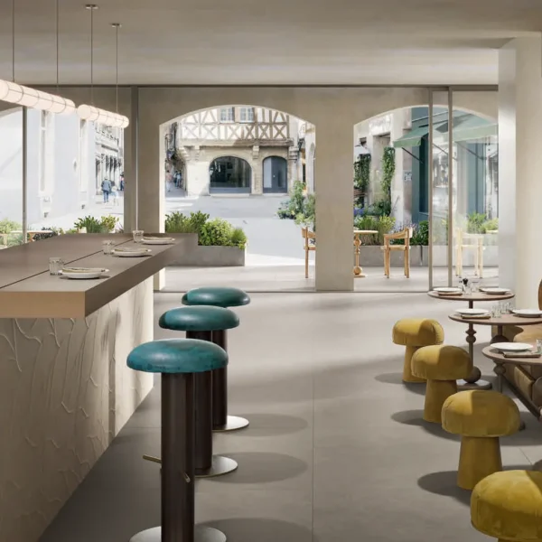 Modern café interior with a long bar, teal stools, and a beige tufted banquette facing large windows overlooking a sunny street.