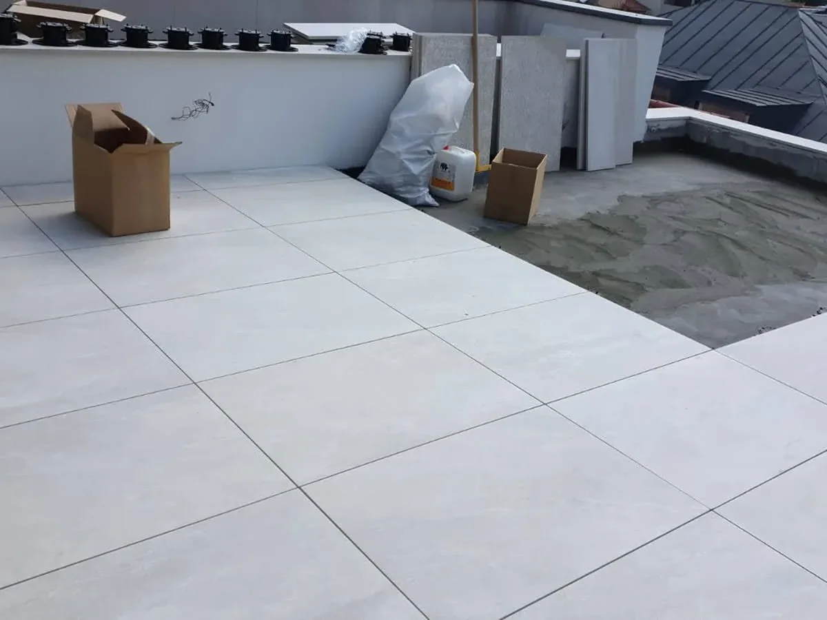 Terasă penthouse București — gresie 2 cm pe ploturi Rooftop terrace under construction with large white tiles installed; cardboard boxes, a white bag, and building materials along the edge.